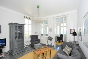 A modern living room with a grey sofa, a wooden coffee table, a grey armchair, a cabinet with glass doors, and a large window.