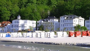 Un hotel frente a la playa con varios edificios blancos, algunos con ventanales salientes y otros con balcones. Hay cabañas de playa blancas y rojas a lo largo de la costa.