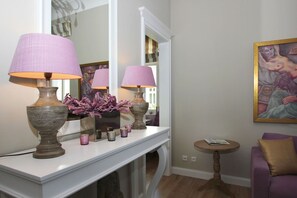 Une pièce avec une console blanche, deux lampes dotées d’abat-jour roses, un vase avec des fleurs violettes, un tableau au mur et un canapé violet.
