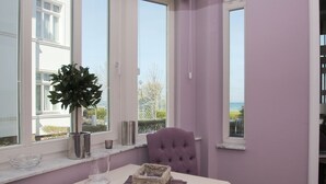Une pièce avec vue sur la mer, une fenêtre ornée d’un pot de fleurs, un fauteuil confortable et une table avec un verre et un panier.