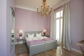 A bedroom with a double bed, two bedside tables with lamps, a chandelier, a large window with curtains, and a mirror on the wall. - Die Ausstattung der Ferienwohnung Nr. 02 in der Villa Strandeck - Villa Strandeck (Binz)