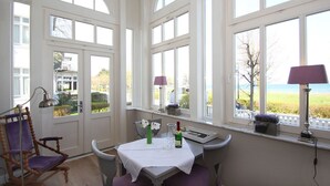 Un coin repas avec une table dressée pour deux personnes, de grandes fenêtres offrant une vue sur la mer et une chaise dotée d’un coussin violet.