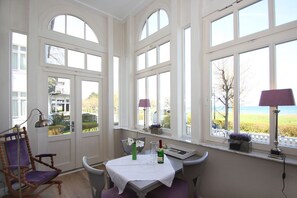 A dining area with a table set for two, large windows with a view of the sea, and a chair with a purple cushion. - Die Ausstattung der Ferienwohnung Nr. 02 in der Villa Strandeck - Villa Strandeck (Binz)