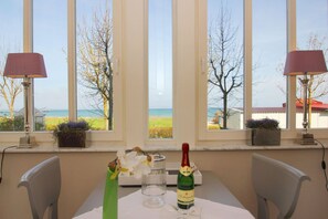 Un coin repas avec une table dressée pour deux personnes, deux lampes, une bouteille de champagne et un vase rempli de fleurs. De grandes fenêtres offrent une vue sur la mer et les arbres.