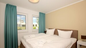 Ein Hotelzimmer mit einem großen Bett, zwei Fenstern mit Vorhängen, einem Nachttisch und einer Lampe.