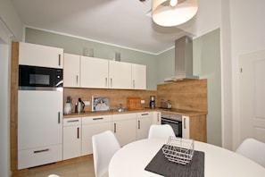 A modern kitchen with white cabinets, a microwave, a coffee maker, and a toaster. There is a dining table with chairs and a small island with a metal rack.