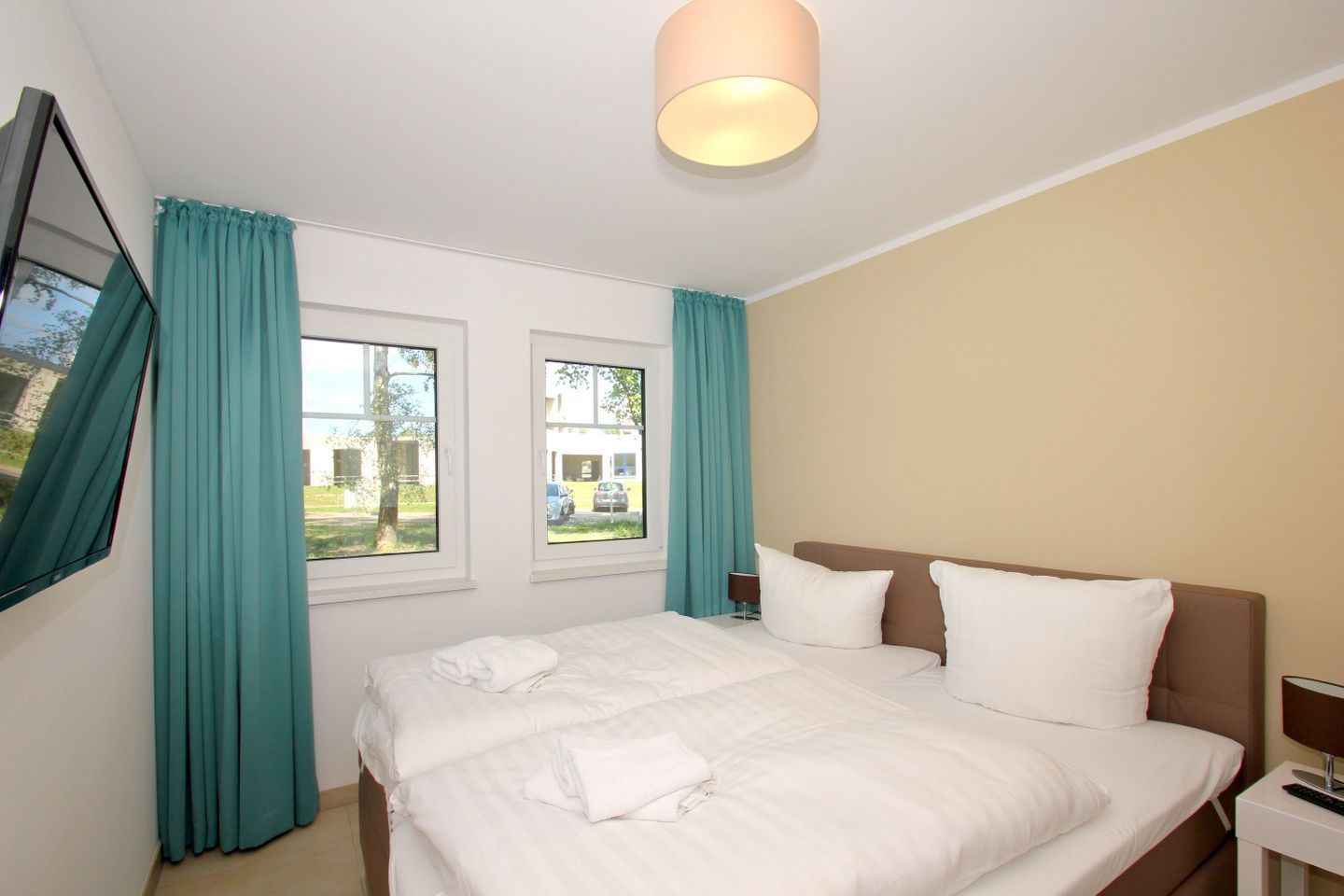 Ein Hotelzimmer mit einem Bett, zwei Fenstern, lindgrünen Vorhängen, einem am Wand montierten Fernseher und einer Deckenleuchte.