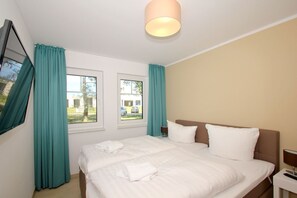 A hotel room with a bed, two windows, teal curtains, a wall-mounted TV, and a ceiling light.