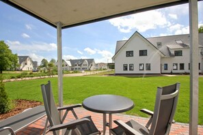 A covered patio with a table and chairs, a lawn, and a white building in the background. - Beachfront vacation rental with a terrace, access to the pool and sauna - Rex Rugia H-1010 - (Göhren)