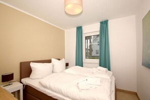 A neatly made bed with white bedding, a bedside table with a lamp, and a window with teal curtains. - Beachfront vacation rental with a terrace, access to the pool and sauna - Rex Rugia H-1010 - (Göhren)