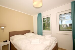 A bedroom with a bed, a nightstand, a lamp, two windows, and a view of the outdoors. - Beachfront apartment with terrace, use of swimming pool and sauna - Rex Rugia apartment 10-2 (Göhren)