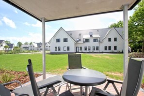 A covered outdoor seating area with a table and chairs, overlooking a grassy lawn and a row of white buildings with dark roofs. - Beachfront apartment with terrace, use of swimming pool and sauna - Rex Rugia apartment 10-2 (Göhren)