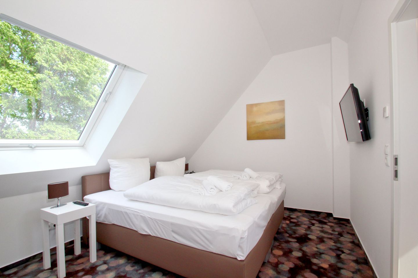 A bedroom with a bed, a small table, a TV, and a painting on the wall.