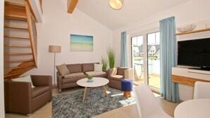 A modern living room with a sofa, coffee table, and TV. There is a staircase on the left and a balcony with a view of houses. - Beachfront vacation rental with a balcony, access to the pool and sauna - Rex Rugia H-1010 - (Göhren)