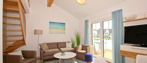 A modern living room with a sofa, coffee table, and TV. There is a staircase on the left and a balcony with a view of houses.