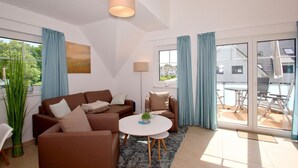 A modern living room with a brown sofa, a small round table, and a floor lamp. There is a large window with blue curtains and a view of a house outside. - Beachfront vacation rental with a balcony, access to the pool and sauna - Rex Rugia H-1010 - (Göhren)
