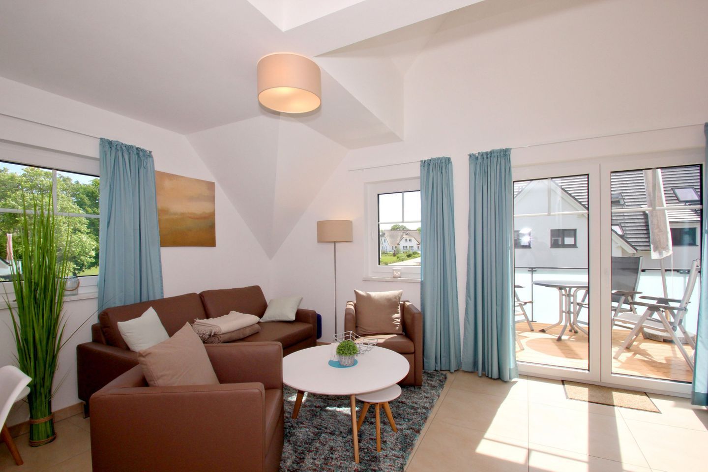 Un salón moderno con un sofá marrón, una pequeña mesa redonda y una lámpara de pie. Hay un ventanal con cortinas azules y vistas a una casa.