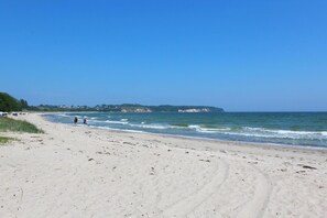 Ein sandiger Strand mit Fußabdrücken, ein klarer, blauer Himmel und eine entfernte Küstenlinie mit Gebäuden.