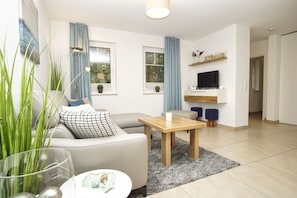 A modern living room with a sofa, a coffee table, and a TV. There are windows with curtains, a plant, and a shelf with decorative items.