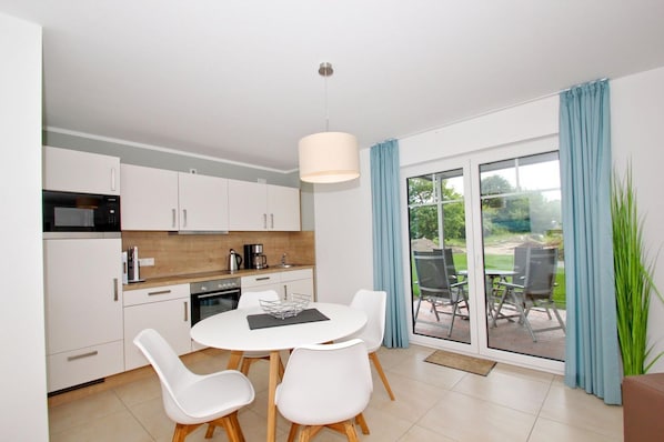 A modern kitchen with white cabinets, a microwave, and a coffee maker. There is a dining area with a white table and chairs, and a view of a garden through large sliding glass doors.