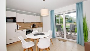 Een moderne keuken met witte kastjes, een magnetron en een koffiezetapparaat. Er is een eethoek met een witte tafel en stoelen, en een uitzicht op een tuin door grote glazen schuifdeuren.