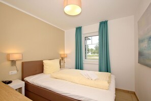 A hotel room with a bed, two bedside lamps, a window with curtains, a painting on the wall, and a wooden nightstand. - Beachfront vacation rental with a terrace, access to the pool and sauna - Rex Rugia - Rex (Göhren)