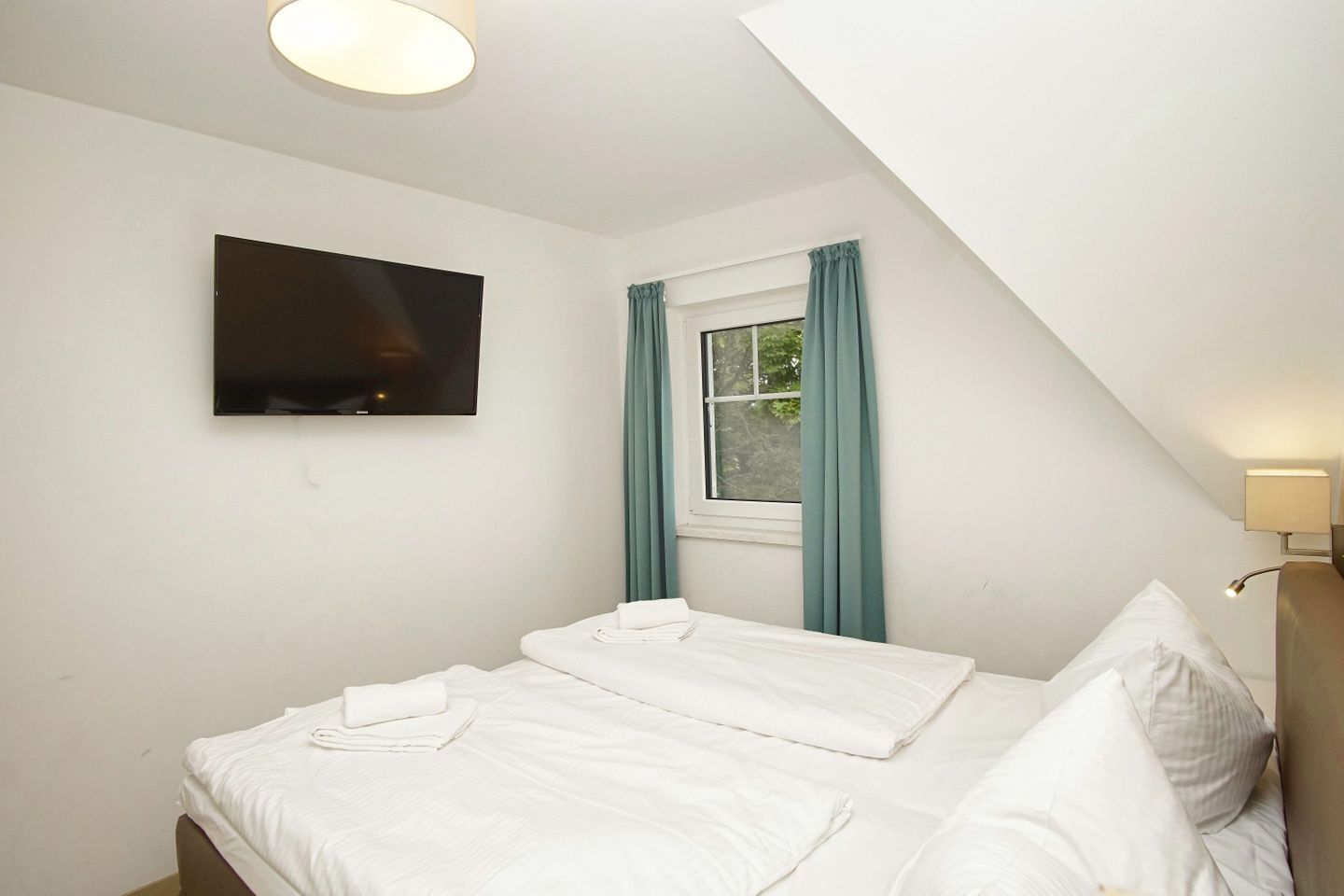 Un dormitorio con cama, televisor, ventana con cortinas y lámpara.