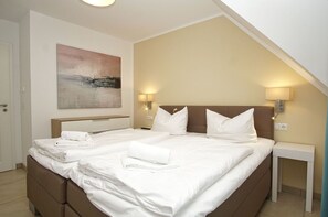A hotel room with a large bed, two bedside tables, a nightstand, a painting on the wall, and a door. - Beachfront apartment with balcony, use of swimming pool and sauna - Rex Rugia H-1010 - (Göhren)