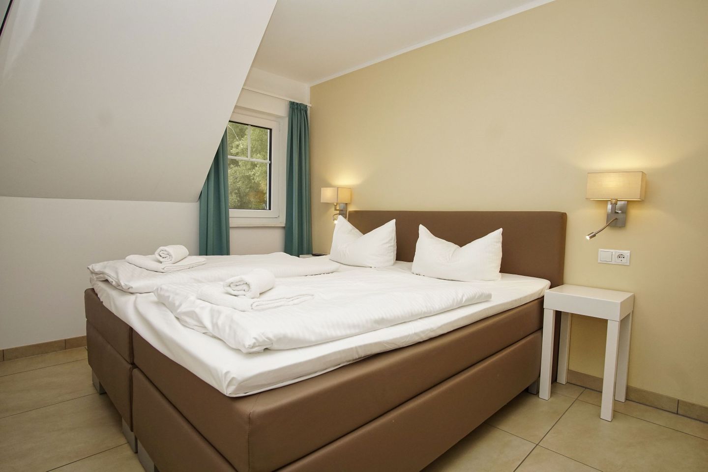 A hotel room with a bed, two bedside lamps, a window with curtains, and a small bedside table.