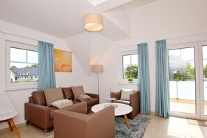 A modern living room with a brown sectional sofa, a white coffee table, and a floor lamp. There are large windows with blue curtains and a view of the outside.