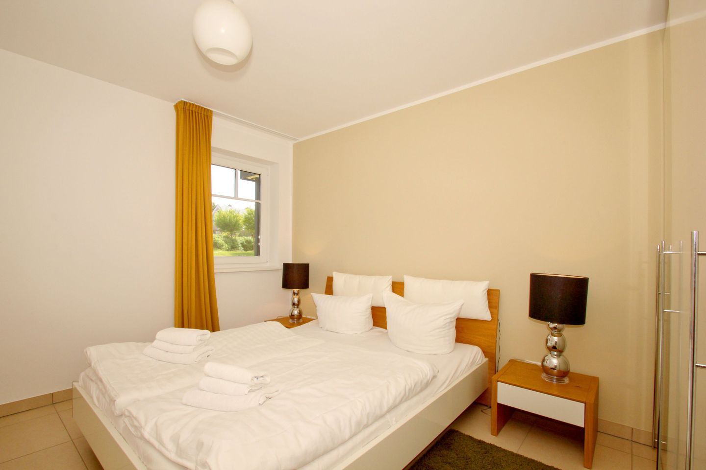 A neatly made bed with white linens and pillows, a bedside table with a lamp, a window with curtains, and a view of greenery outside.