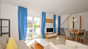 Ein modernes Wohnzimmer mit Sofa, Fernseher, Esstisch, Stühlen und einem Balkon mit blauen Vorhängen.