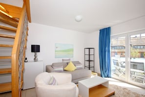 A modern living room with a staircase, a sofa, a coffee table, a lamp, a painting, and a view of the city through large windows. - Beachfront apartment with a balcony; access to the pool and sauna - Rex Rugia Apartment 28-6 - (Göhren)