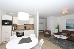 A modern kitchen with white cabinets, a dining table with chairs, a sofa, a coffee table, and a painting on the wall. - Beachfront vacation rental with a terrace, access to the pool and sauna - Rex Rugia - Rex (Göhren)