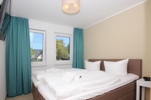 A hotel room with a bed, two windows, teal curtains, a bedside table, and a ceiling light.