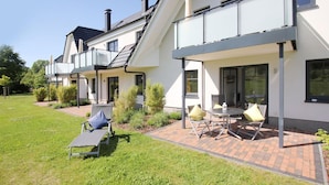 Ein modernes, zweistöckiges Haus mit einer Terrasse, die Gartenmöbel und einen kleinen Tisch bietet.