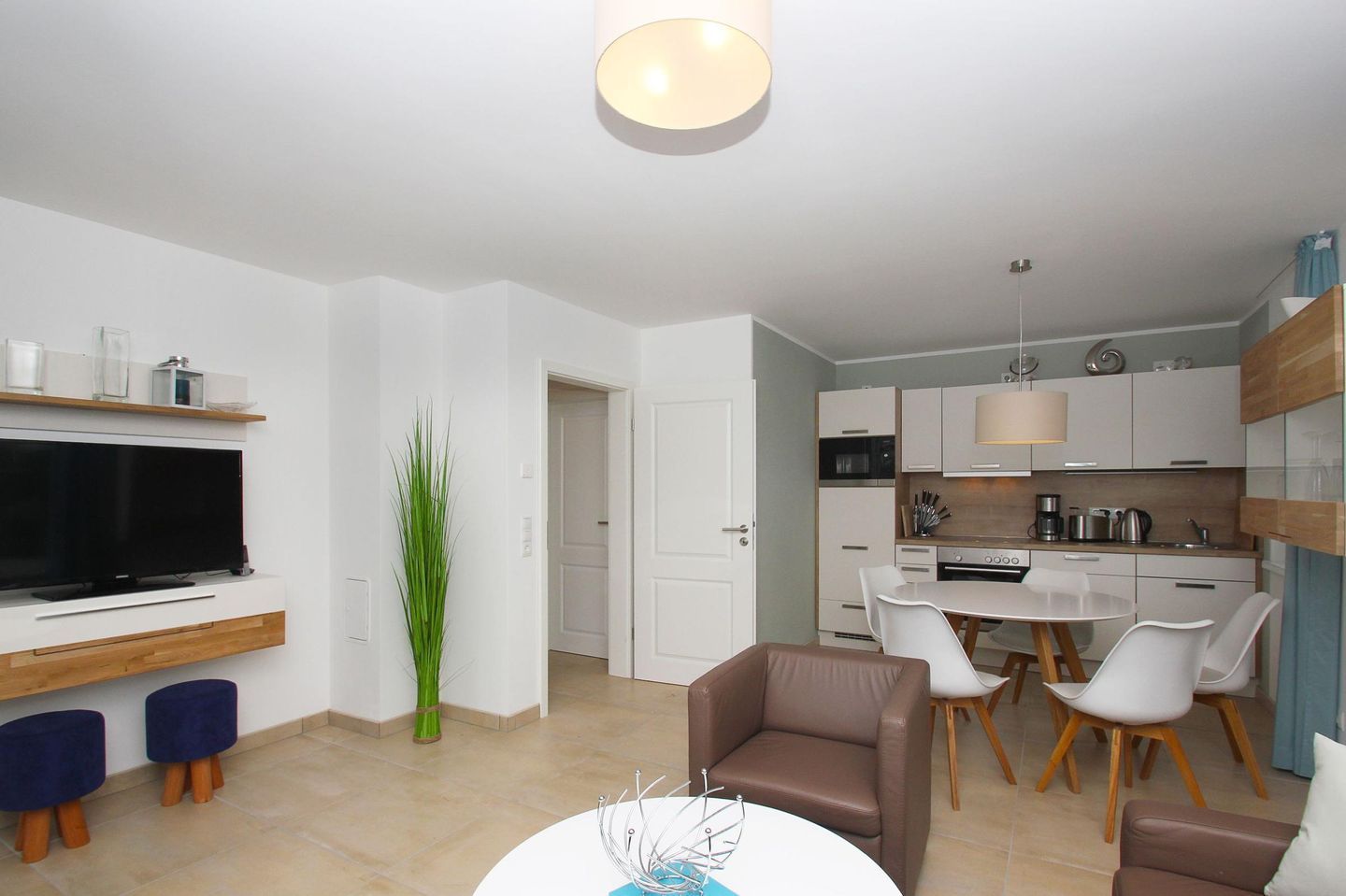 Una sala de estar moderna con un televisor de pantalla plana, una zona de comedor con mesa y sillas blancas, y una cocina con gabinetes blancos y encimera de madera.