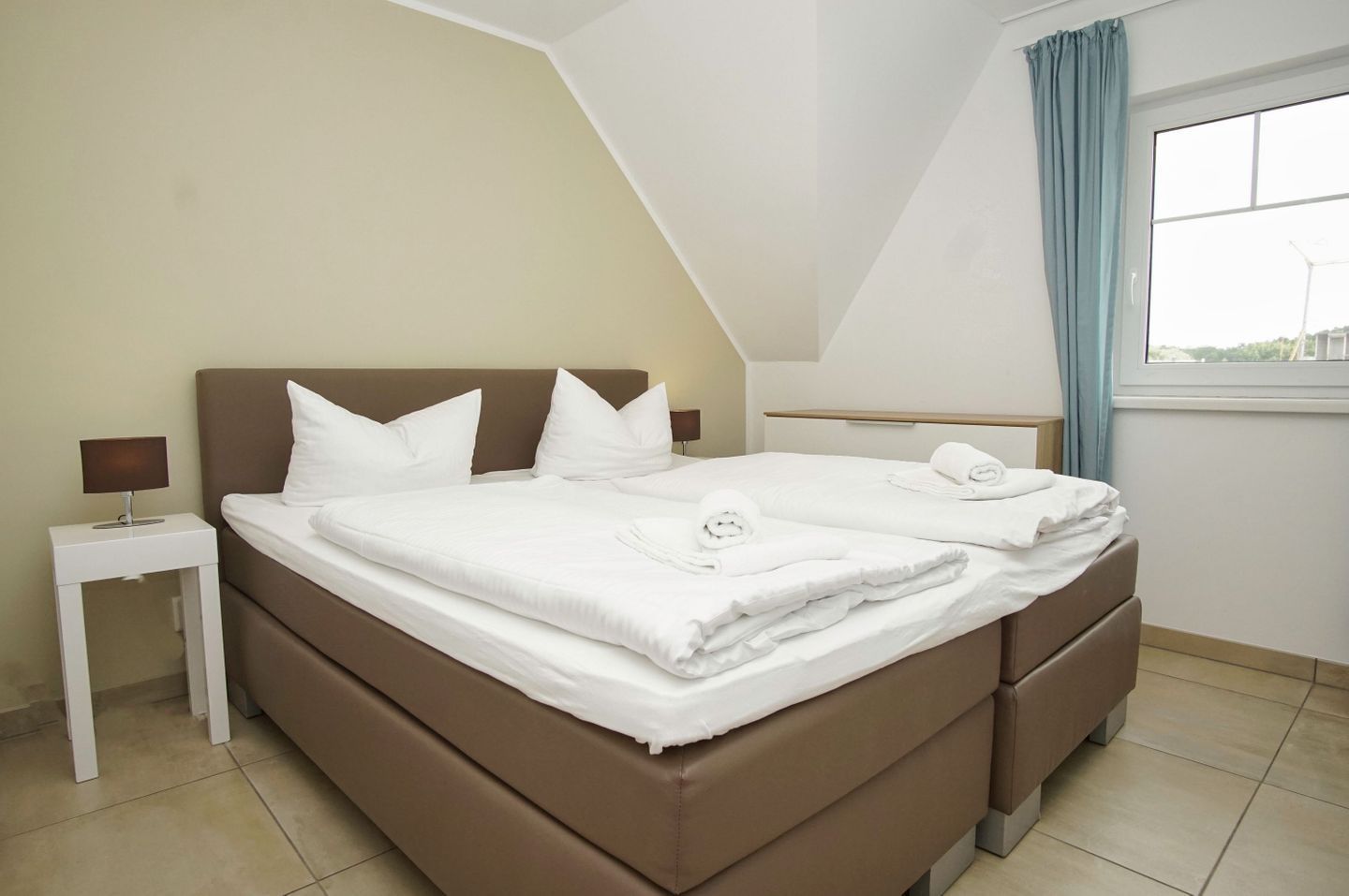 Een slaapkamer met een bruin bed, witte kussens, een nachtkastje met een lamp en een raam met gordijnen.