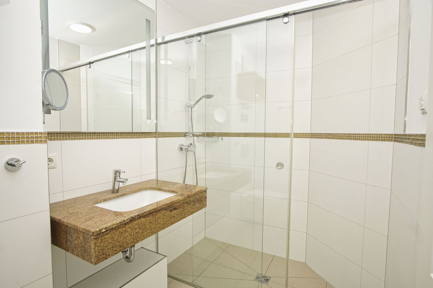Un baño moderno con ducha envuelta en vidrio, lavamanos de encimera de granito y paredes con azulejos blancos.