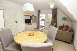 A modern living room with a round wooden table, grey upholstered chairs, a beige sofa, a flat-screen TV, and a balcony with a view.