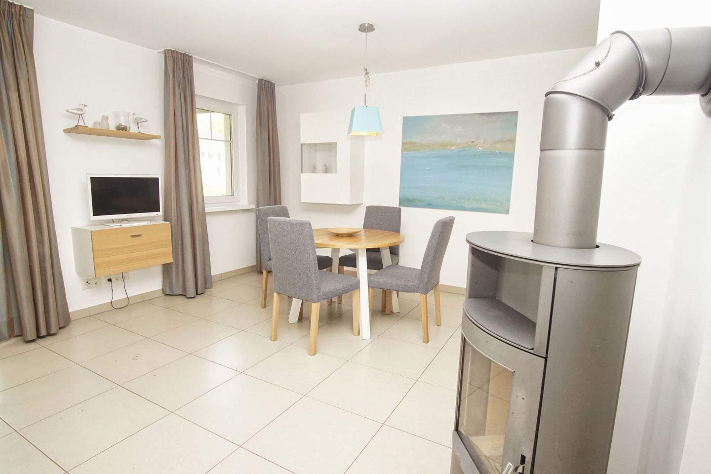 A modern living room with a fireplace, a dining table with chairs, a TV mounted on the wall, and a painting of a seascape.