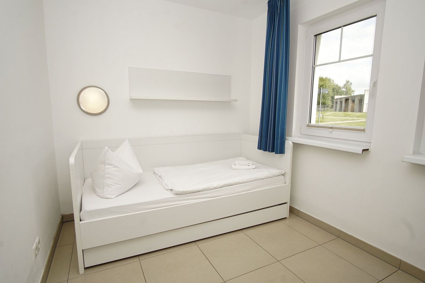 A white room with a bed, a window with blue curtains, and a wall-mounted light fixture.