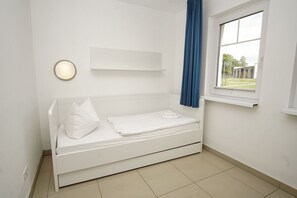A white room with a bed, a window with blue curtains, and a wall-mounted light fixture. - Beachfront vacation rental with a terrace, access to the pool and sauna - Rex Rugia - Rex (Göhren)