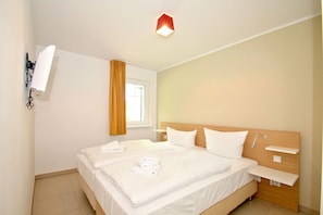 A hotel room with a bed, a bedside table, a wall-mounted TV, a window with curtains, and a ceiling light. - Beachfront vacation rental with a terrace, access to the pool and sauna - Rex Rugia - Rex (Göhren)