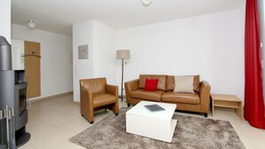 A modern living room with a sofa, armchair, and a coffee table. There is a lamp, a clock, and a red curtain. - Beachfront vacation rental with a terrace, access to the pool and sauna - Rex Rugia - Rex (Göhren)
