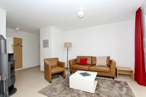 Un salon moderne avec un canapé, un fauteuil et une table basse. On y trouve une lampe, une horloge et un rideau rouge.