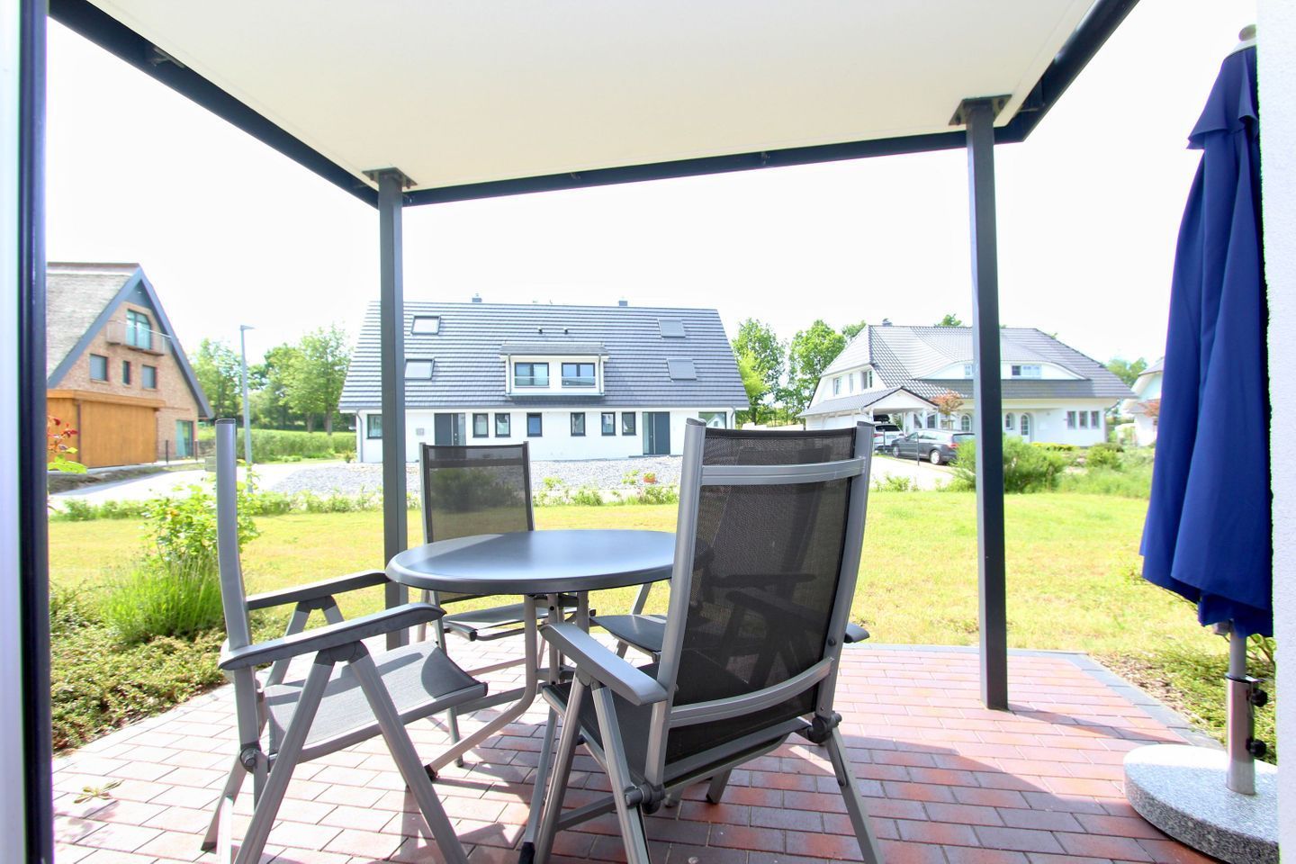 Eine Terrasse im Freien mit Tisch und Stühlen, einem blauen Sonnenschirm und Blick auf Häuser im Hintergrund.