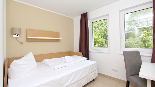 A hotel room with a bed, a desk, a chair, a wall-mounted lamp, and a window with curtains. - Beachfront vacation rental with a terrace, access to the pool and sauna - Rex Rugia - Rex (Göhren)