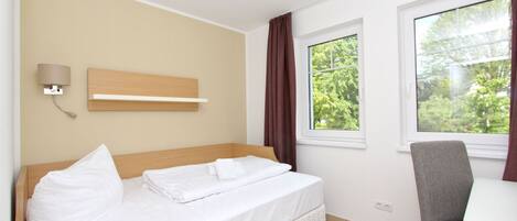 A hotel room with a bed, a desk, a chair, a wall-mounted lamp, and a window with curtains.