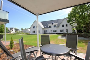 An outdoor patio with a table and chairs, a covered area, and a view of a large white building with multiple windows and a sloped roof.